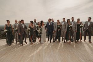 Full wedding party lined up on wooden deck for formal group photo under overcast sky.