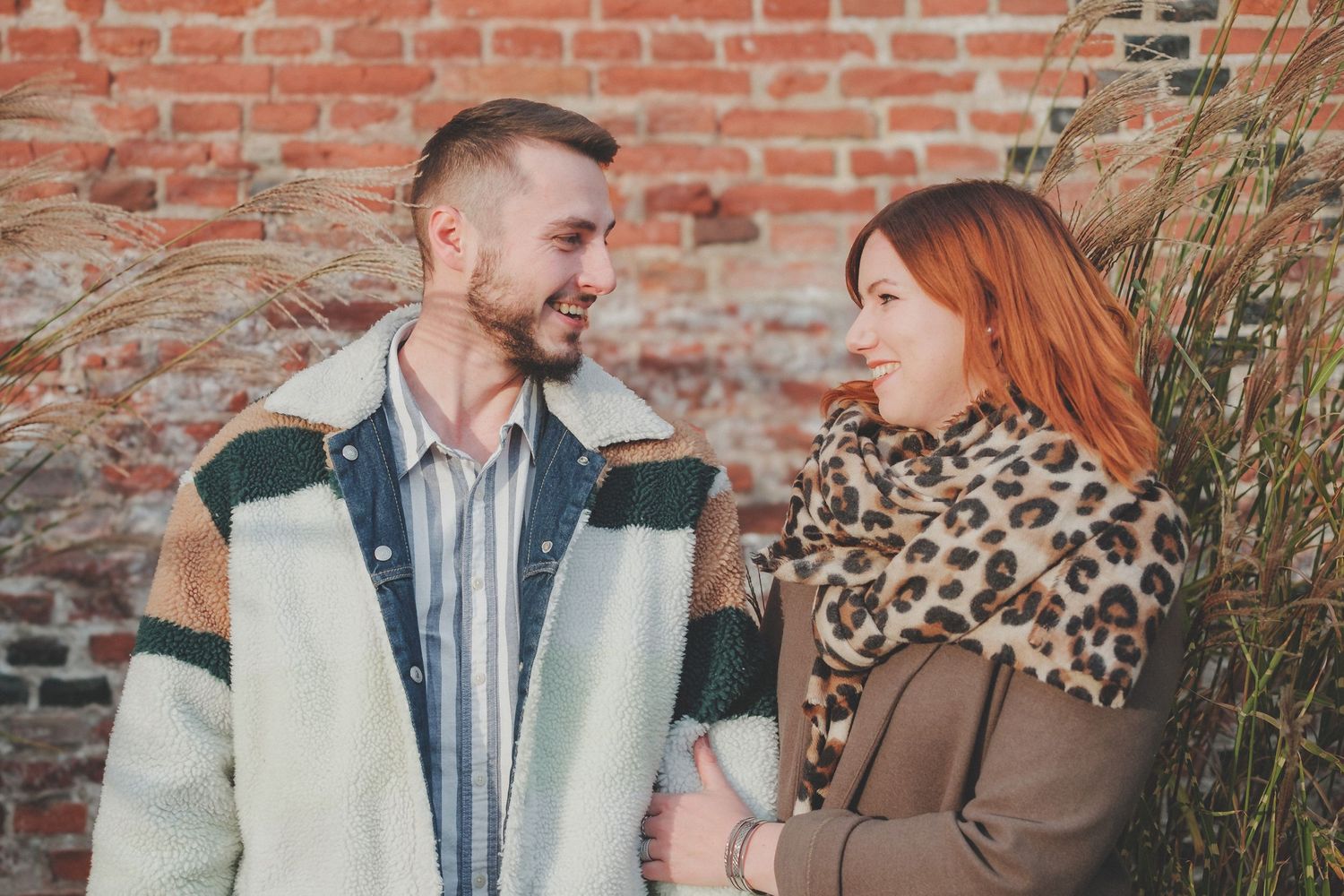 Couple in casual winter wear laughing together against a rustic brick wall background.