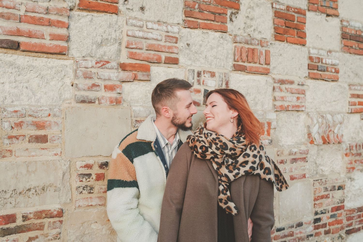 A couple in winter clothing stands against a weathered brick wall sharing a tender moment.