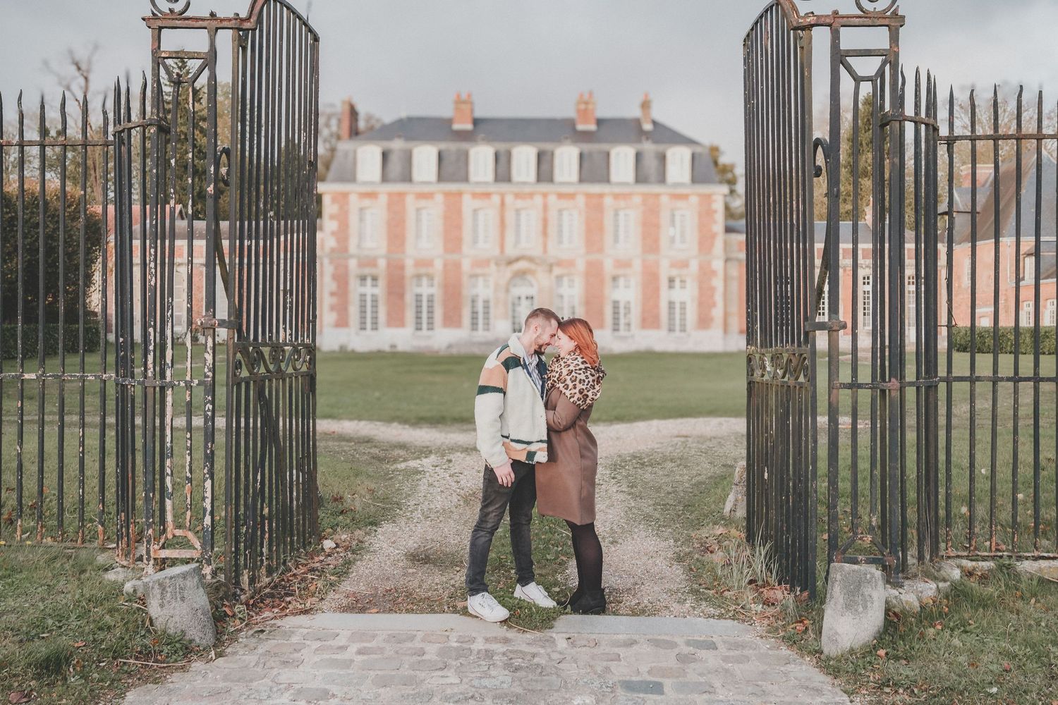 A romantic couple stands between ornate gates with a grand mansion in the background.
