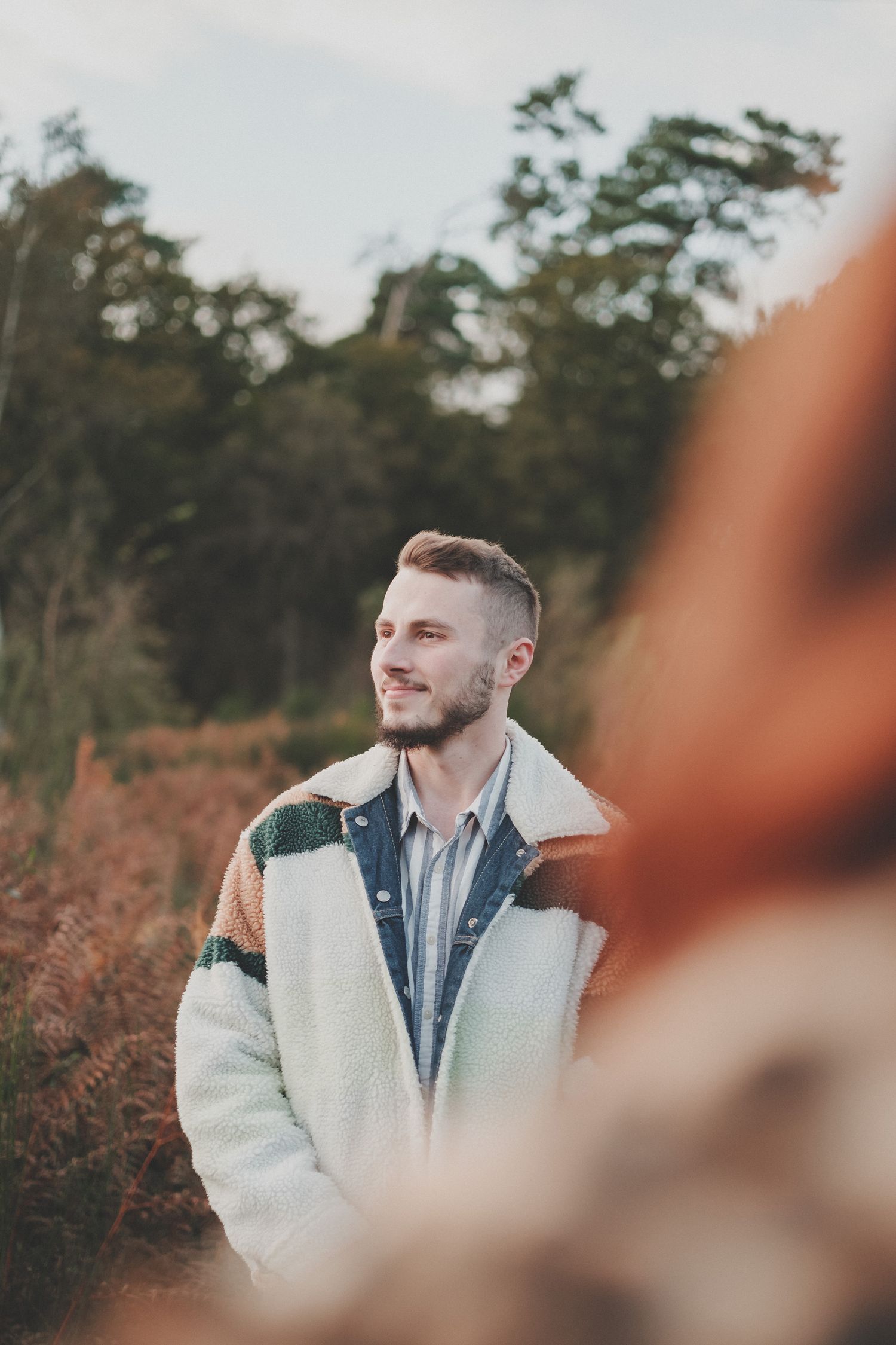 A person in a color-block sherpa jacket smiles while standing in an autumn forest setting.