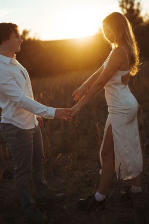 Couple holding hands against golden sunset in field wearing white formal attire.