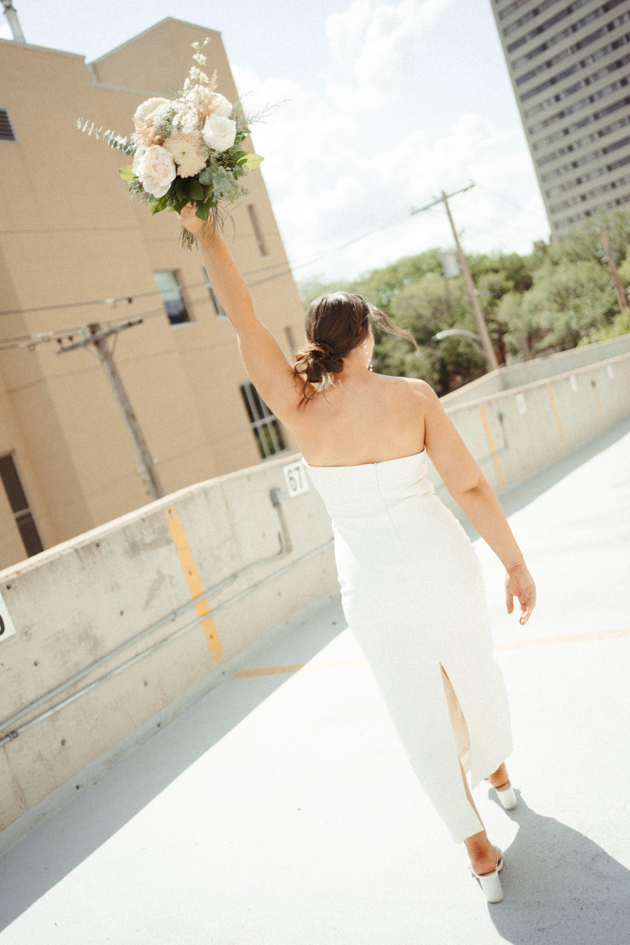 Bride in fitted white dress raises bouquet triumphantly on urban rooftop.