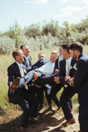 Groomsmen in dark suits lifting and carrying someone while laughing outdoors.