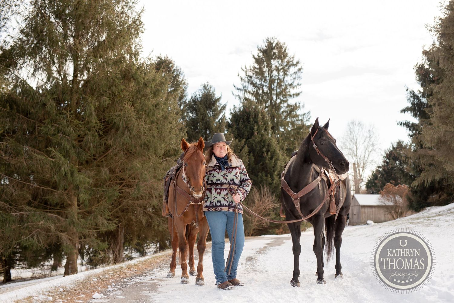 Two horses and their handler walk along a snowy path with evergreen trees in the background.