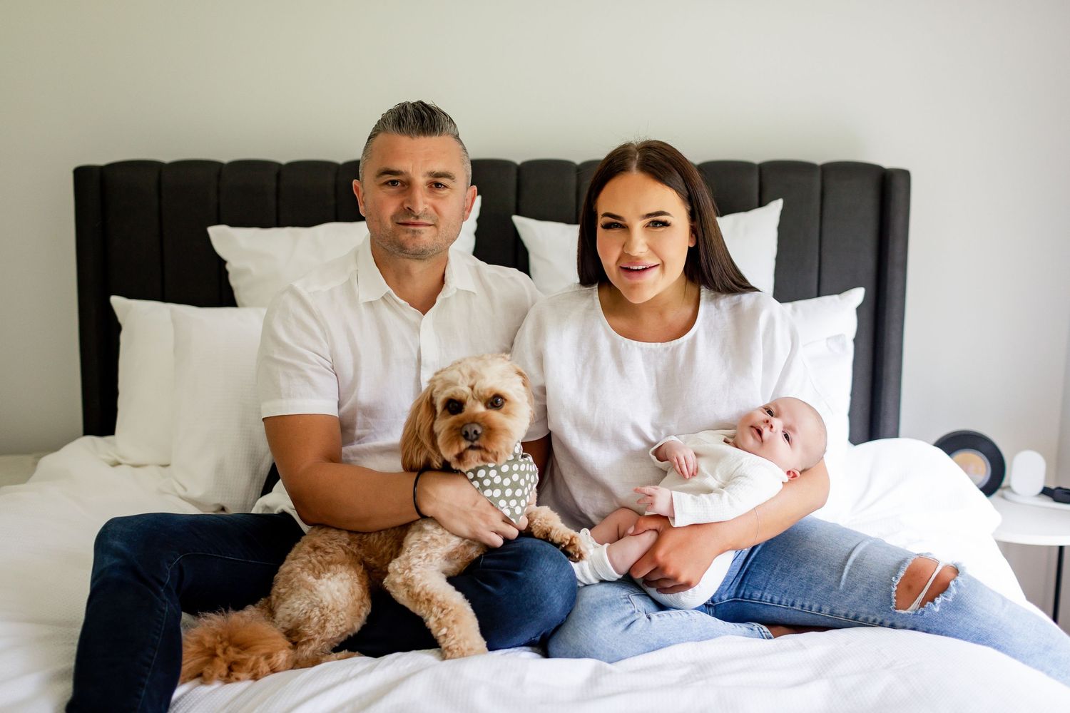 Happy couple sits on bed with their small dog and baby in matching white outfits.