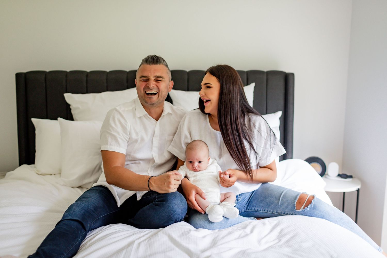 A couple sits on a bed laughing while holding their infant baby in a bright bedroom.