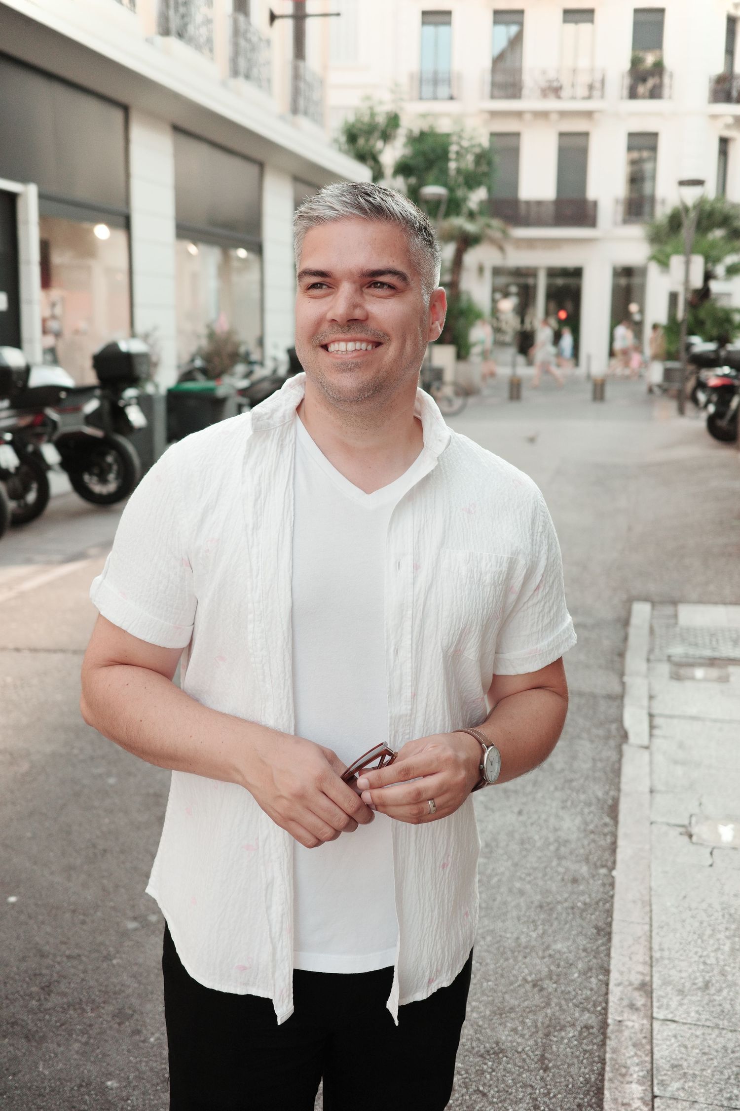 Person in white shirt smiling on urban street with buildings and motorcycles.