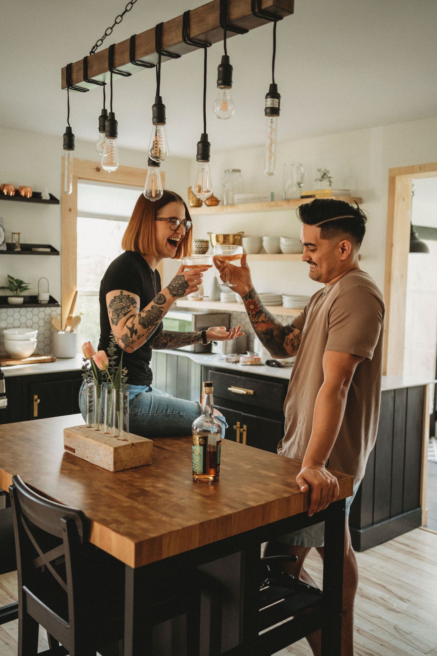 Couple toasting drinks in modern kitchen with industrial lighting fixtures.