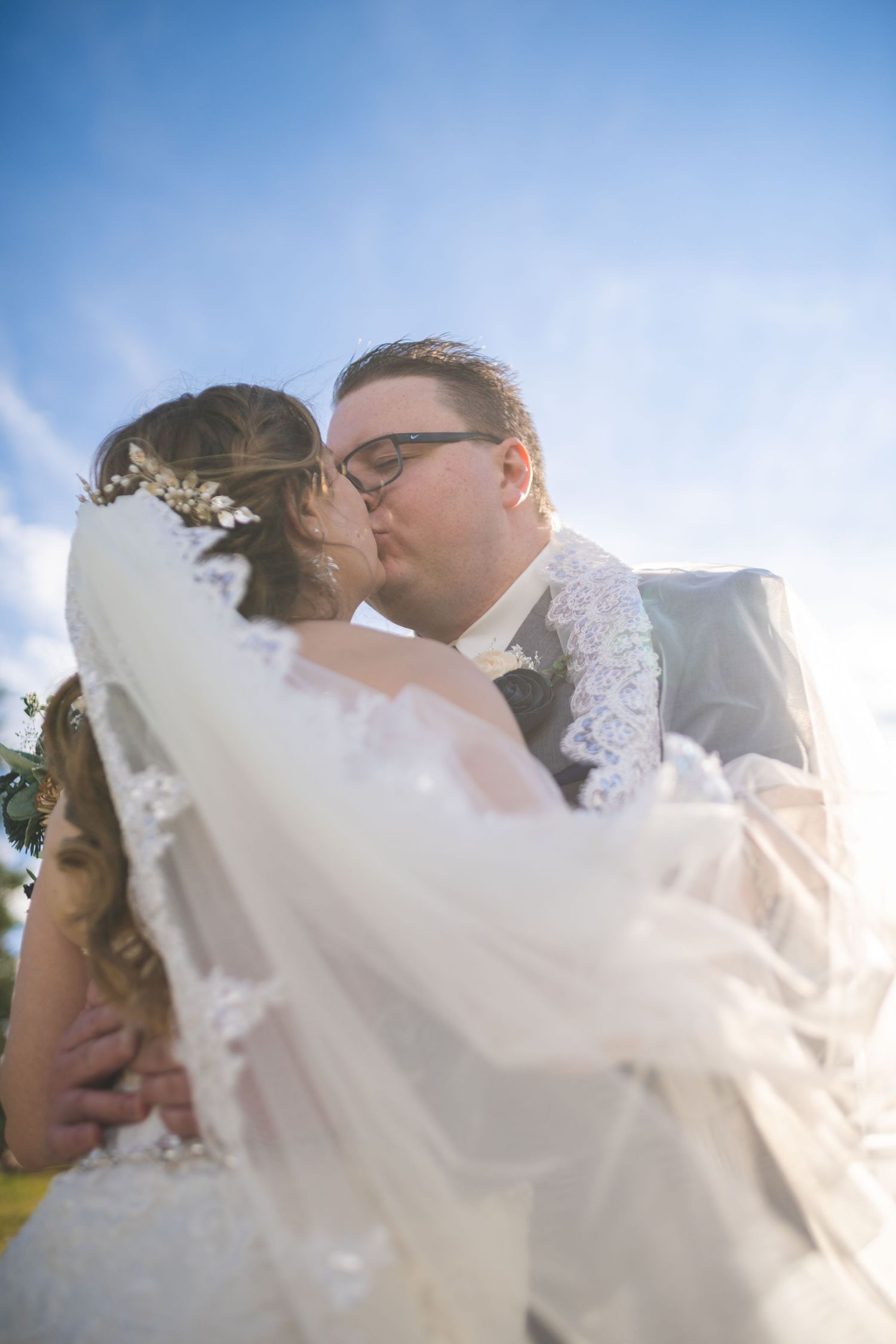 looking up at a bride with long veil kissing her groom who is wearing glasses