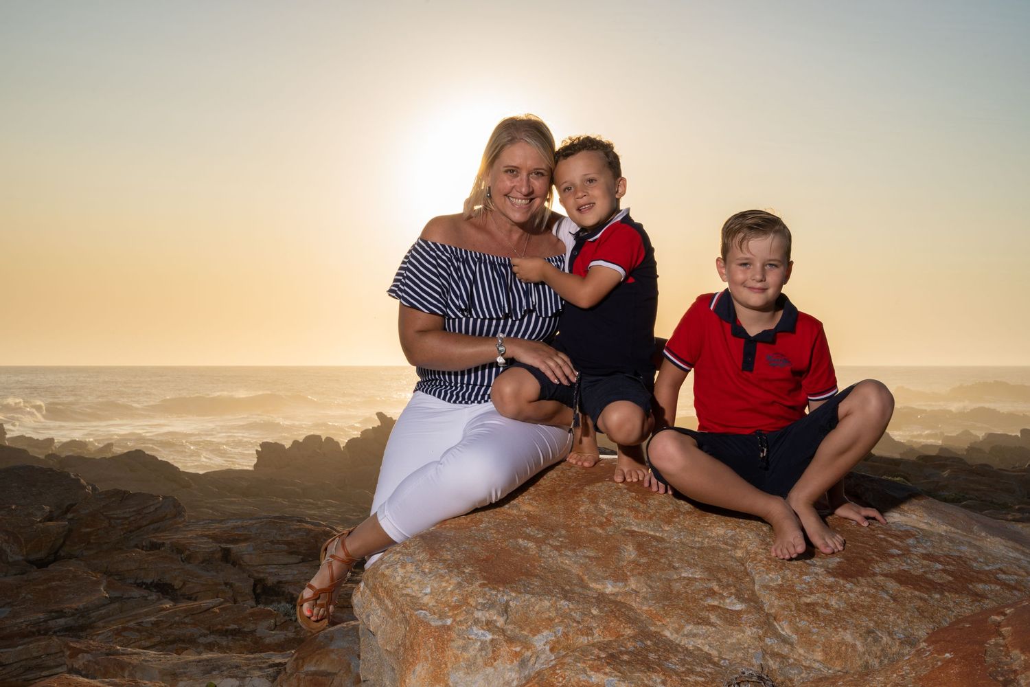 A mother and her two young sons pose on rocky shore at golden sunset by the ocean.