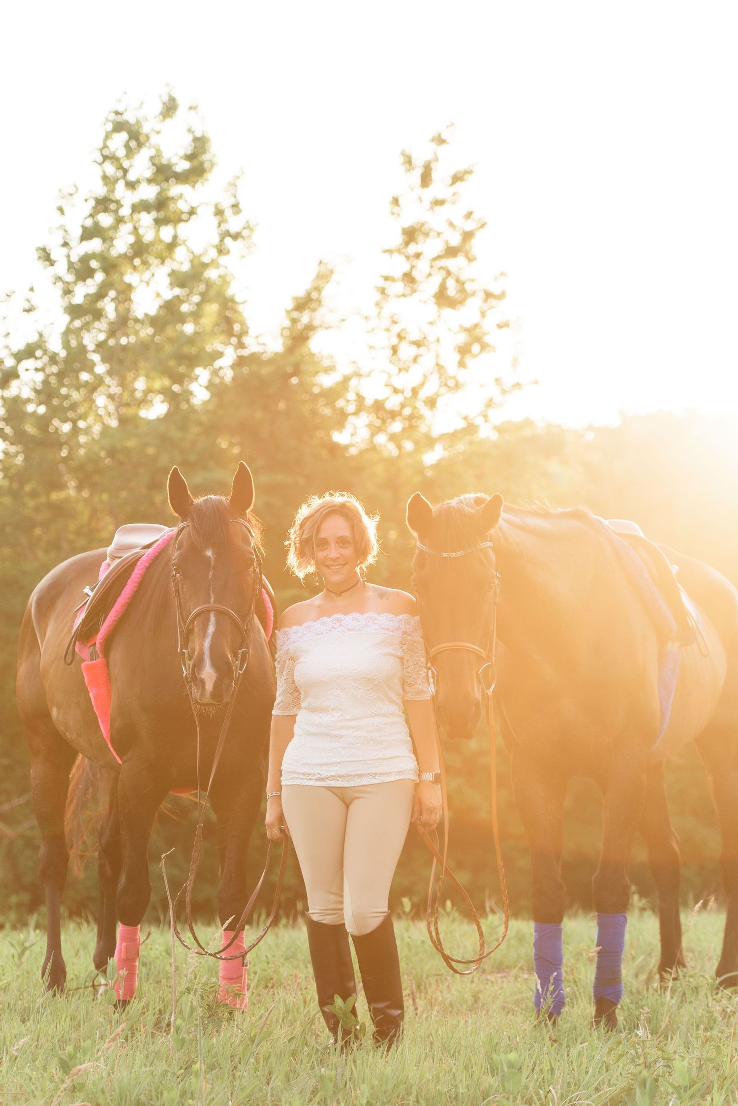 Woman in white top and riding breeches standing between two dark horses in golden sunlight.