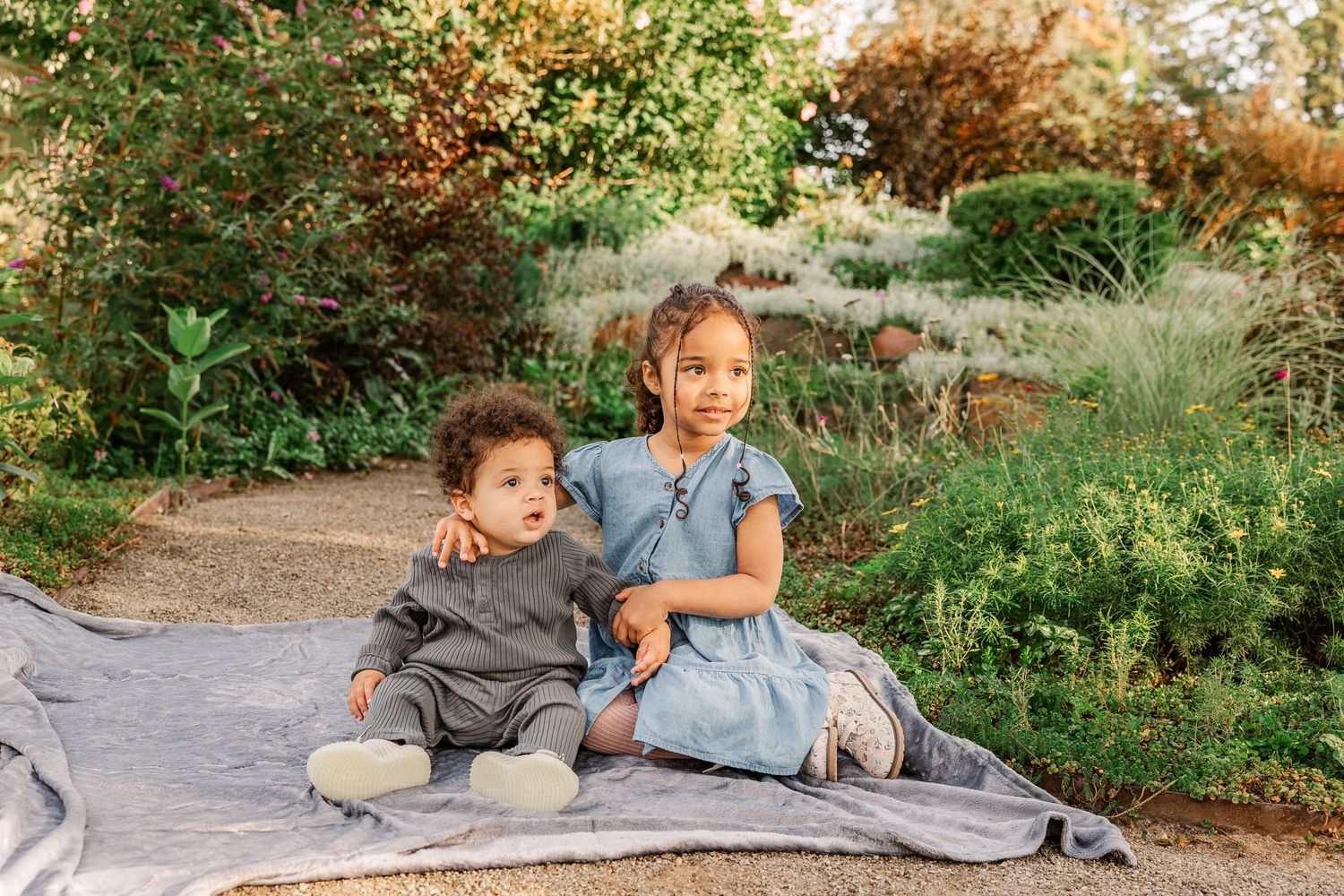 Children share a sweet moment while sitting on a gray blanket in a garden.