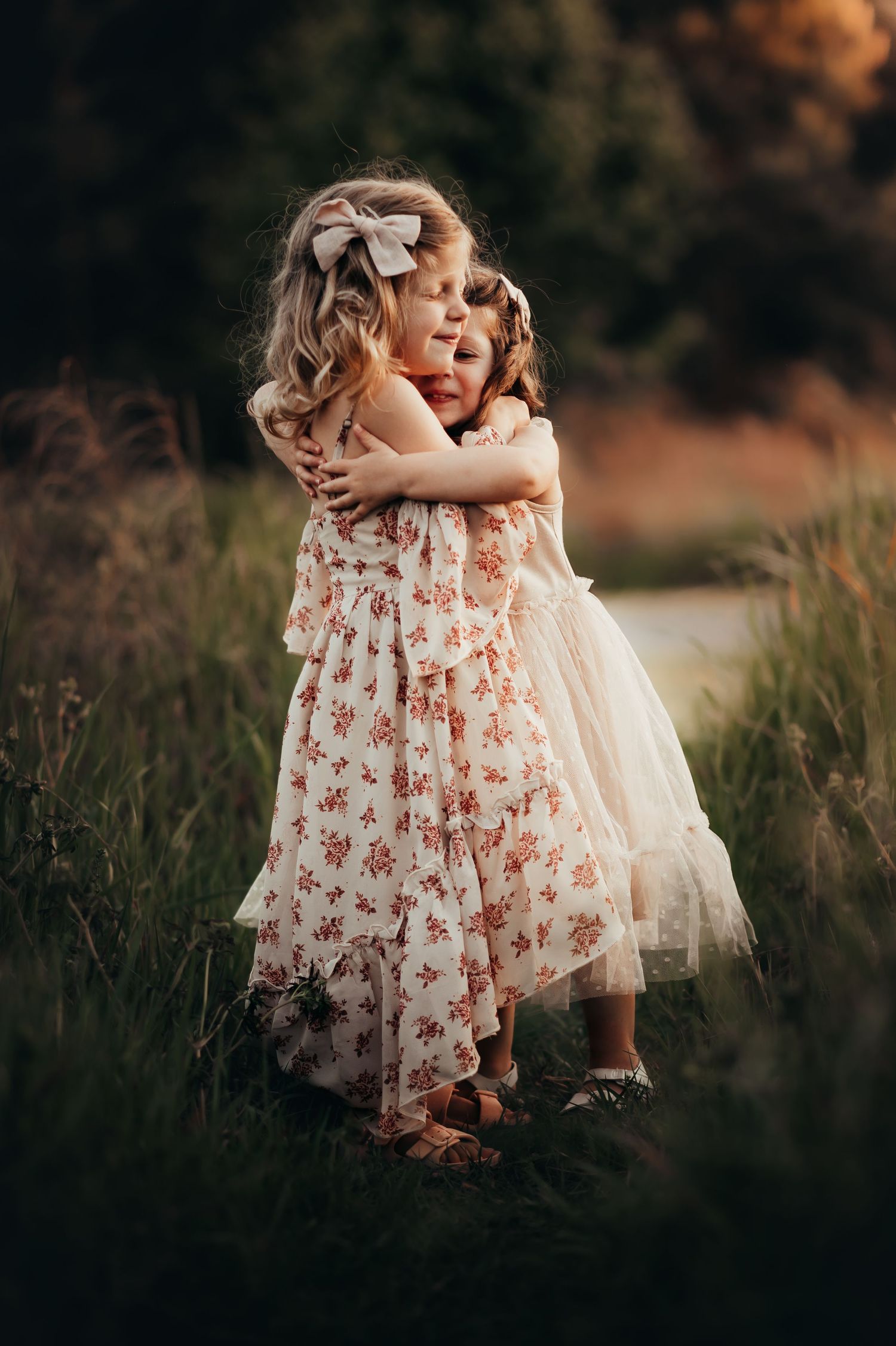 Younger cousin hugs her 1-year-older blonde cousin in tall grass at sunset. Both girls wear Joyfolie dresses and bows.