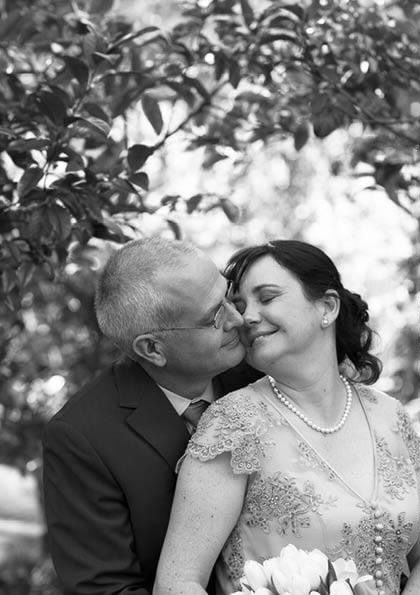 Black and white wedding photo of a couple sharing a tender moment outdoors, bride in lace dress with pearl necklace holding flowers.