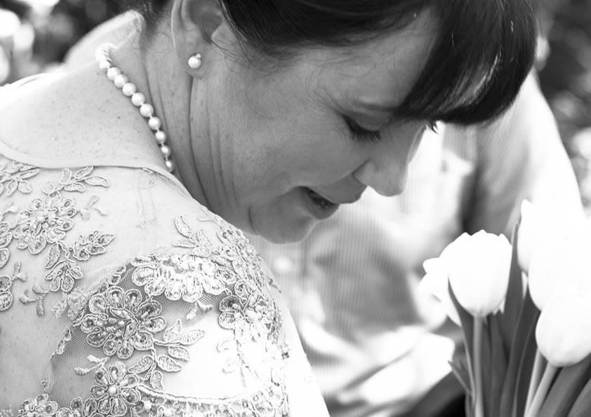 Black and white photo of a bride in lace gown wearing pearl necklace, looking down at white tulips.