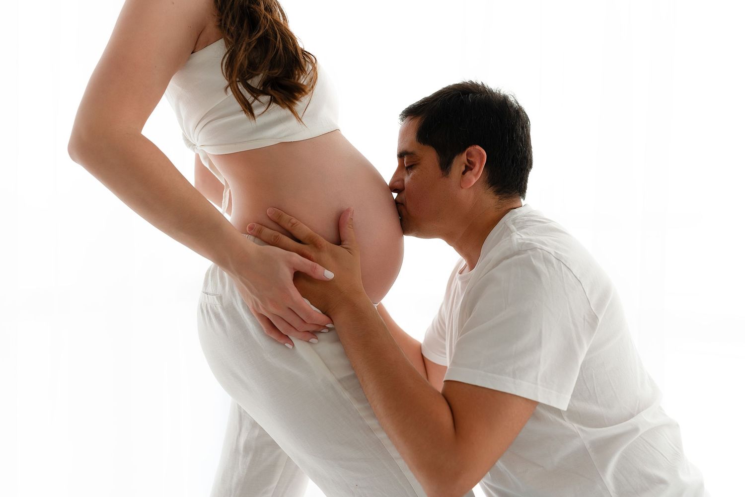 A couple embraces during intimate maternity photoshoot against white backdrop.