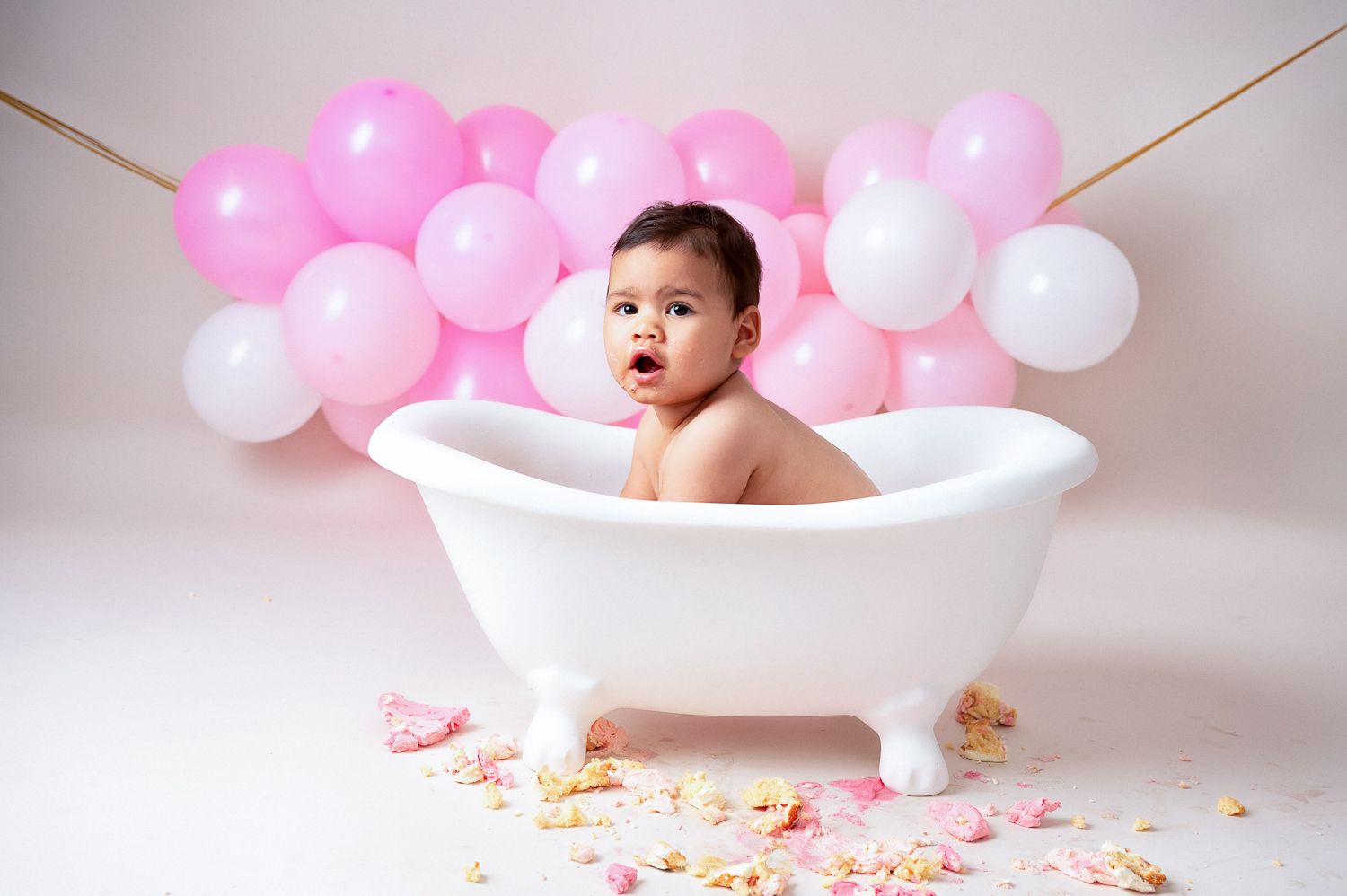 Baby in white bathtub with pink and white balloons backdrop.