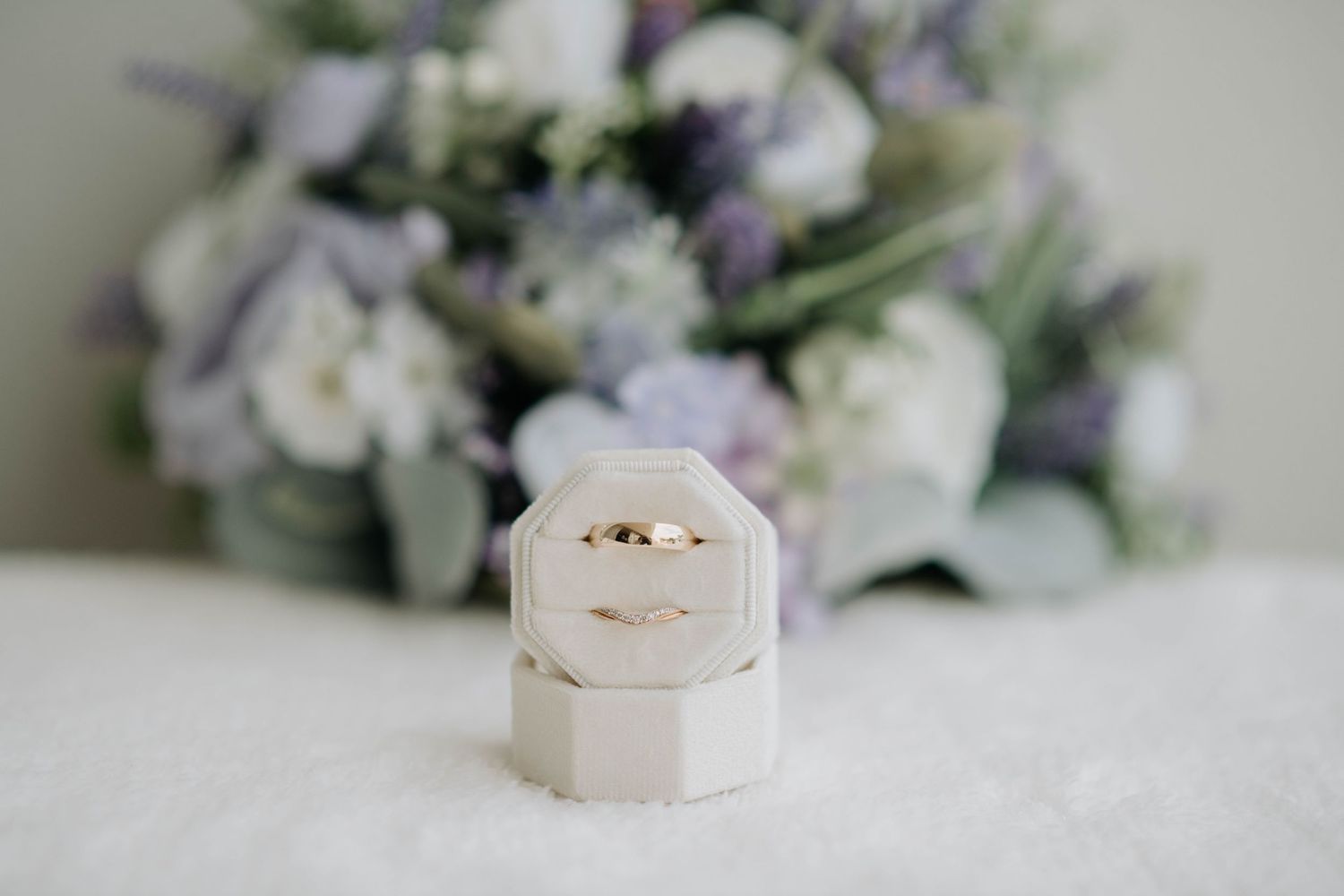 Wedding rings displayed in hexagonal white box with purple and white floral arrangement.