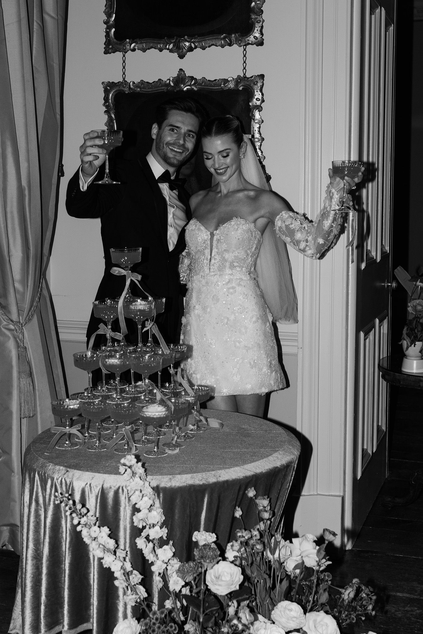 Couple celebrates by champagne tower at wedding reception in vintage-style black and white photo.