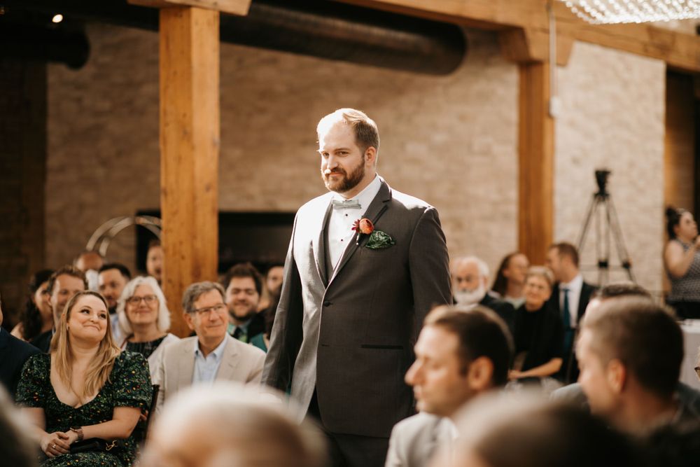Groom in a dark suit with boutonniere smiling as he walks down the wedding aisle.