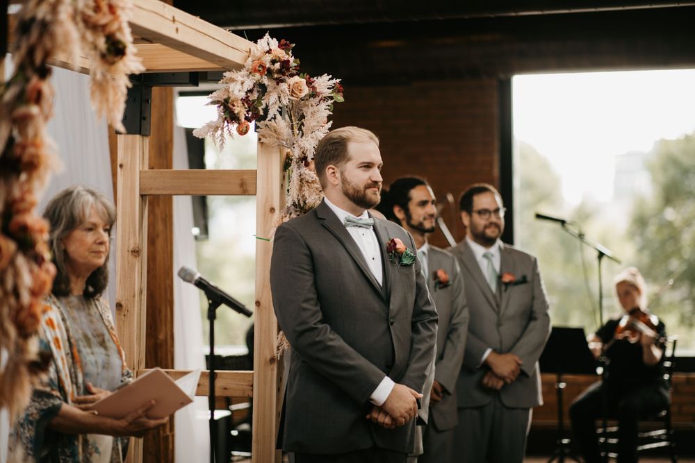 Groom and groomsmen standing at the altar beneath a floral wooden arch during ceremony.
