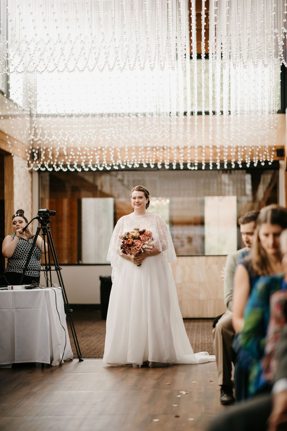 Smiling bride in white tulle gown holding bouquet walks down aisle under crystal chandelier at indoor wedding ceremony.