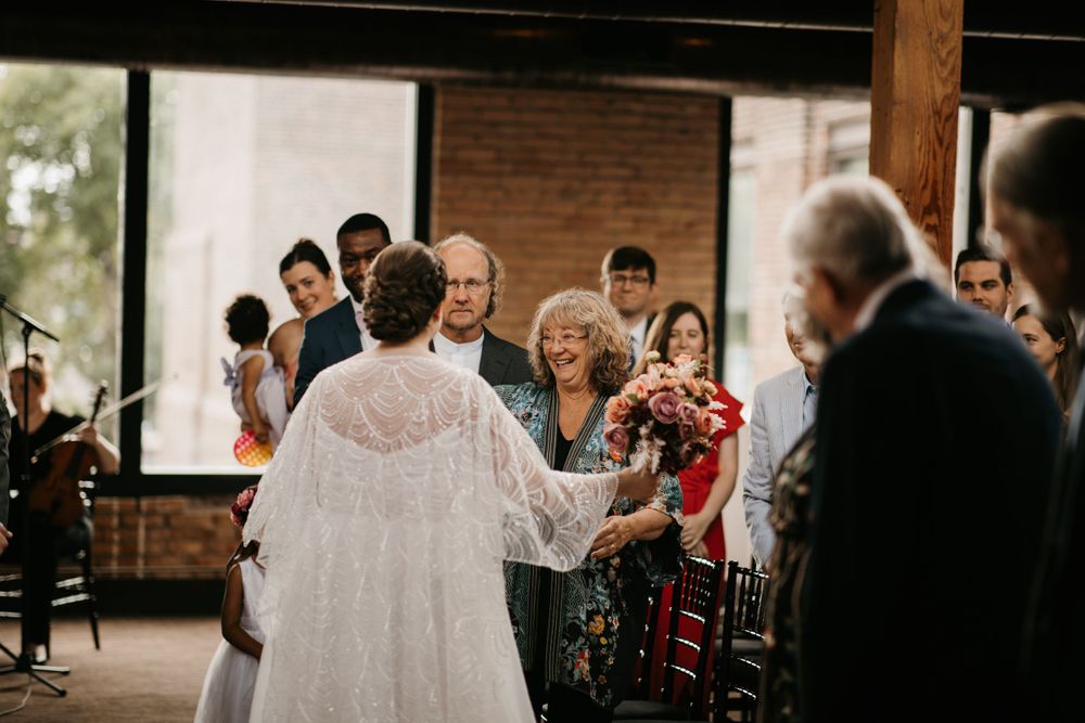 Bride in lace cape gown greets smiling guests while walking down the aisle at an indoor wedding ceremony.