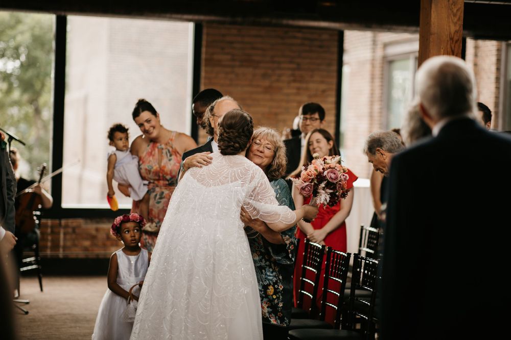 Bride in lace wedding gown embraces elderly guest while walking down the aisle at an indoor wedding ceremony.