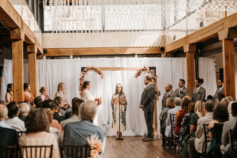 Bride and groom stand at floral arch altar surrounded by wedding party during indoor ceremony with crystal chandelier.