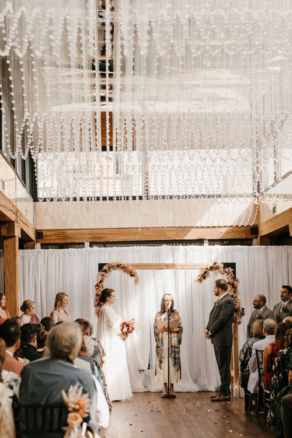 Bride and groom exchange vows under floral arch with crystal chandelier overhead at elegant indoor wedding ceremony.