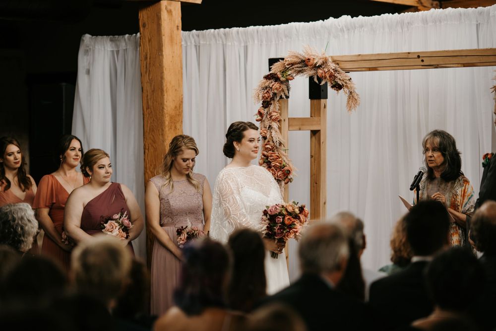 Bride holding autumn bouquet stands at altar with bridesmaids in dusty rose gowns during indoor wedding ceremony.