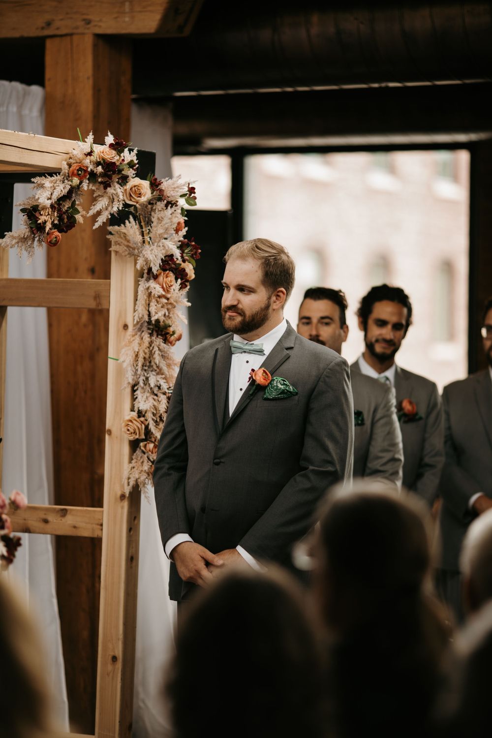 Emotional groom in gray suit with orange boutonniere waits at floral arch altar during indoor wedding ceremony.