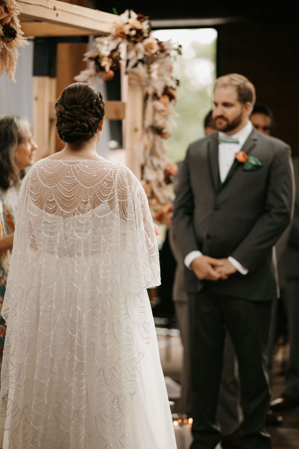 Bride in intricate lace cape gown faces groom in gray suit at rustic wooden arch during wedding ceremony.