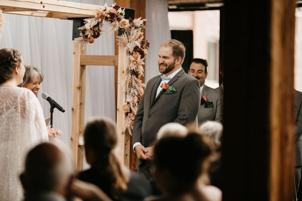 Groom in gray suit smiles warmly at bride during indoor wedding ceremony beside rustic floral wooden arch.