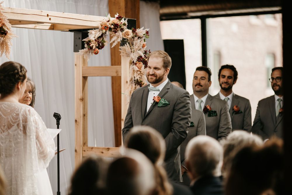 Smiling groom in gray suit stands at floral wooden arch altar with groomsmen during indoor wedding ceremony.