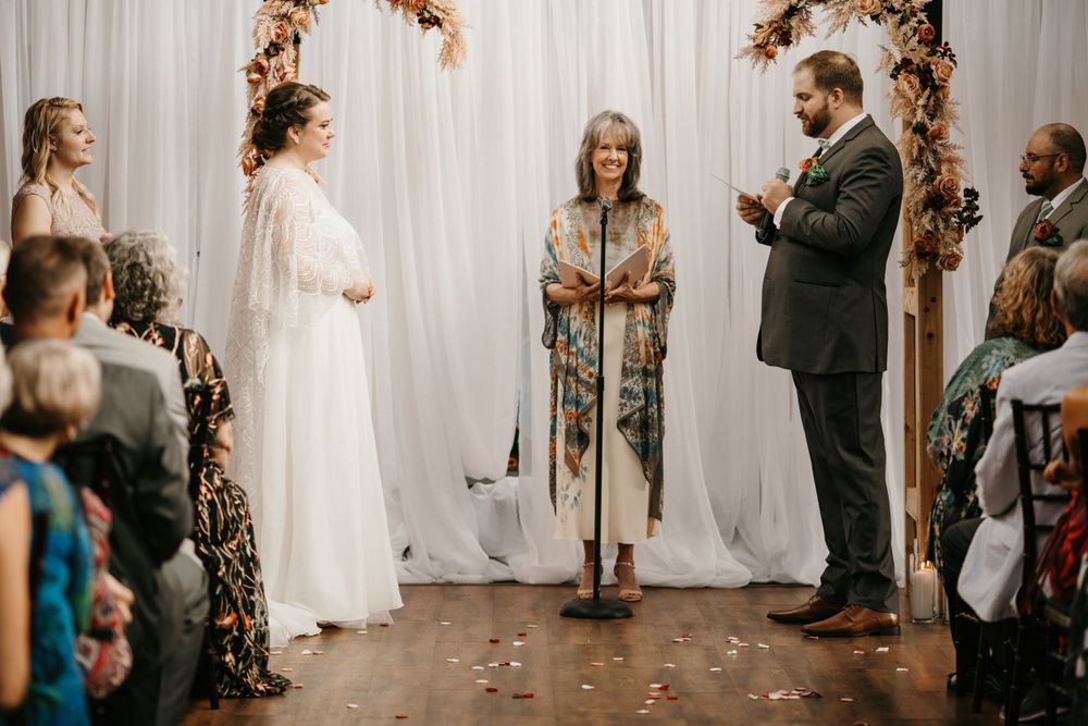 Bride and groom stand at a floral arch altar as an officiant reads during an indoor wedding ceremony.