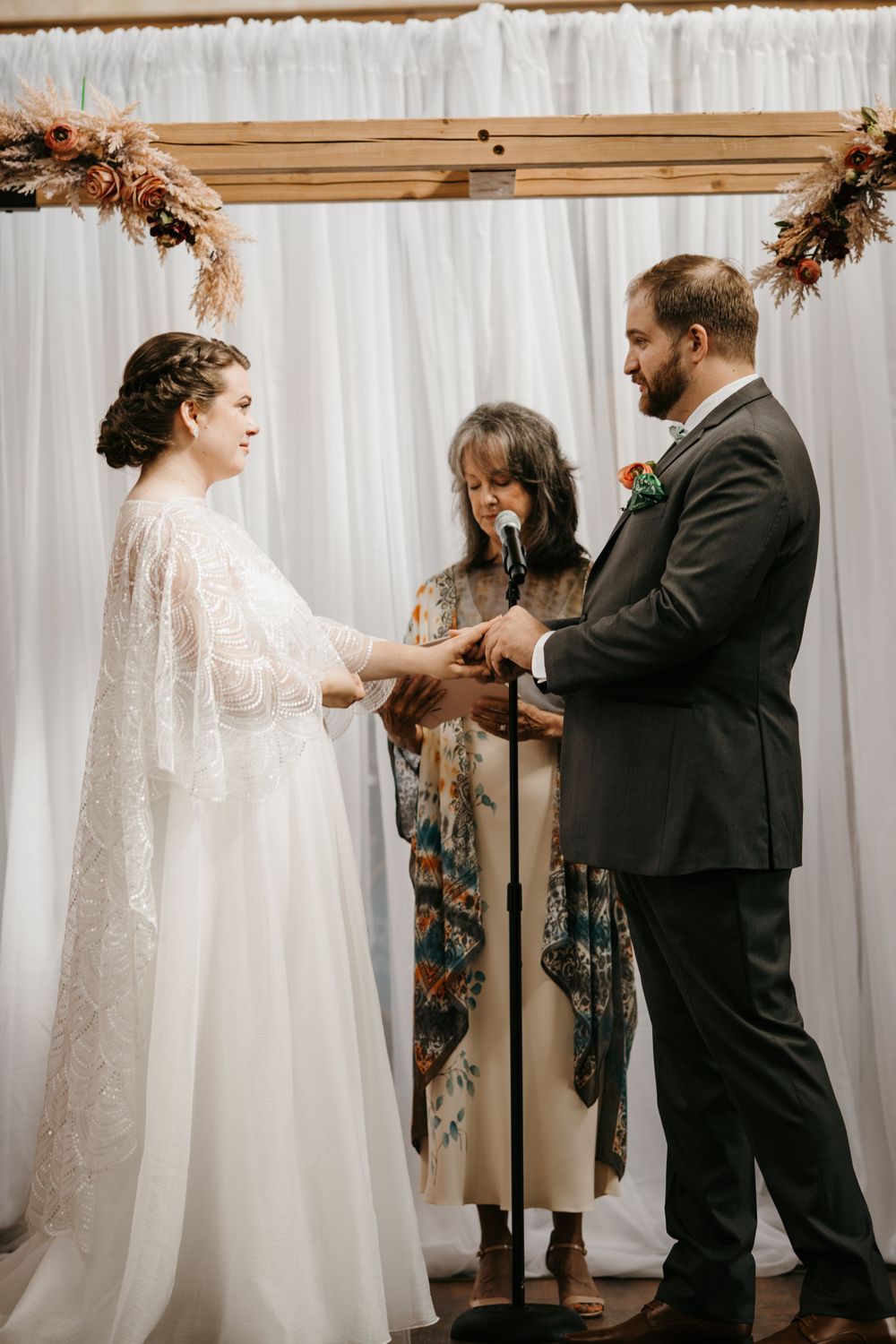 Groom places ring on bride's finger at a wooden arch altar with dried floral arrangements indoors.