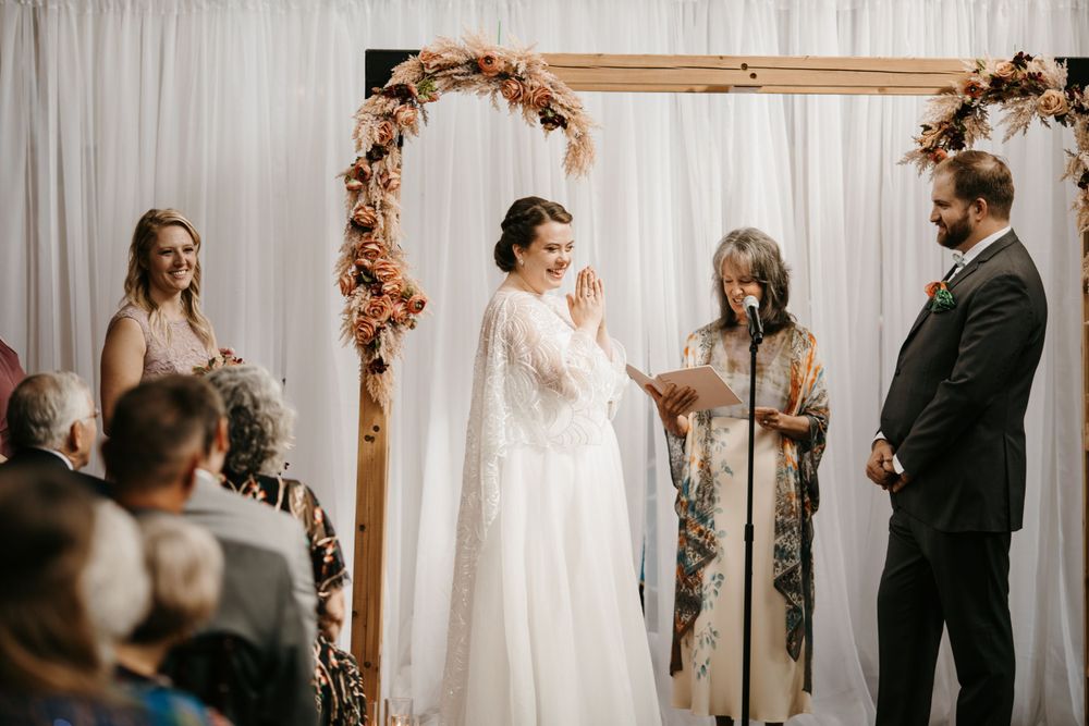 Bride clasps hands joyfully at a rustic wooden arch altar during an indoor wedding ceremony.
