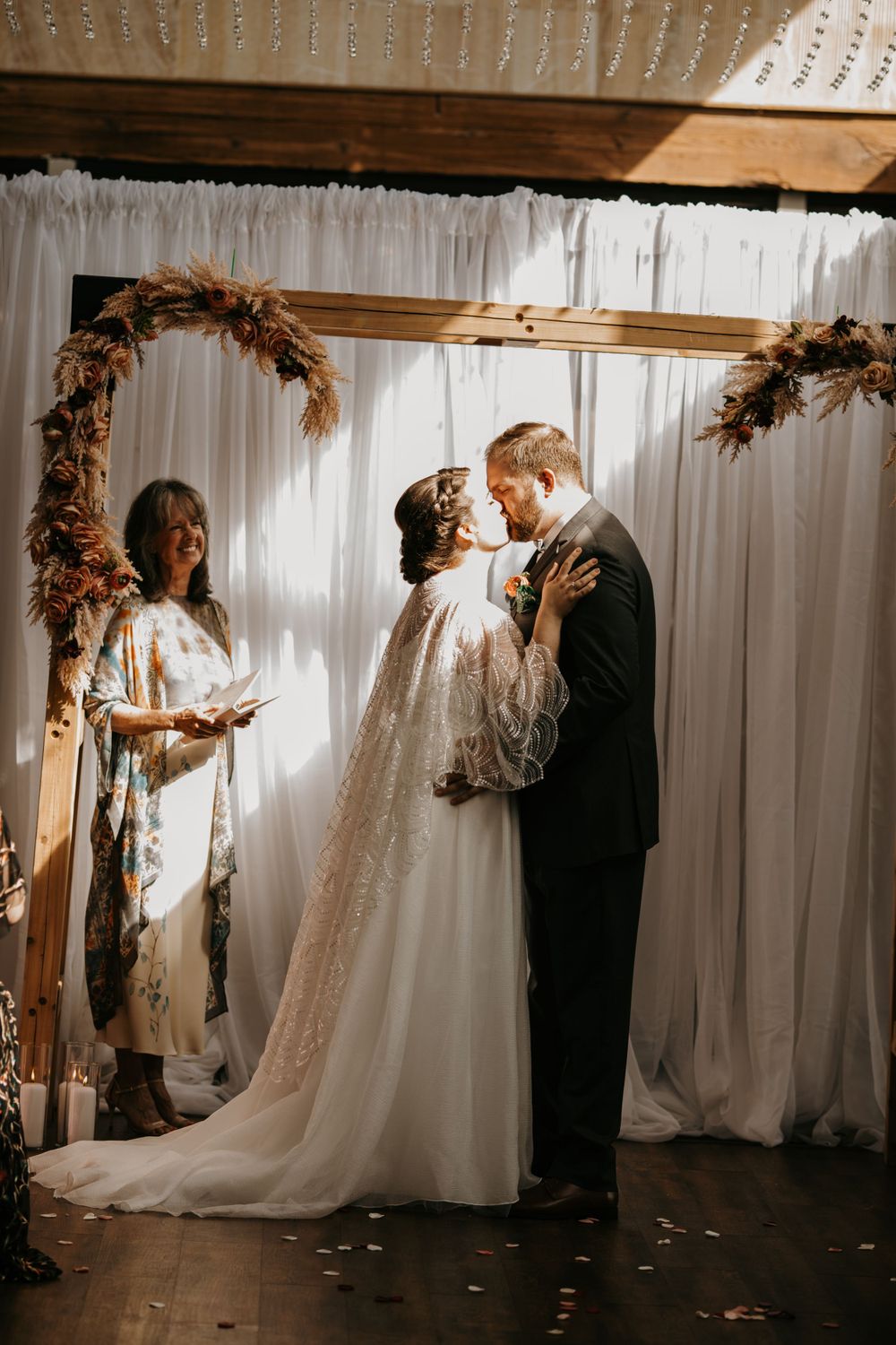Bride and groom share their first kiss under a dried floral wooden arch at an indoor wedding.