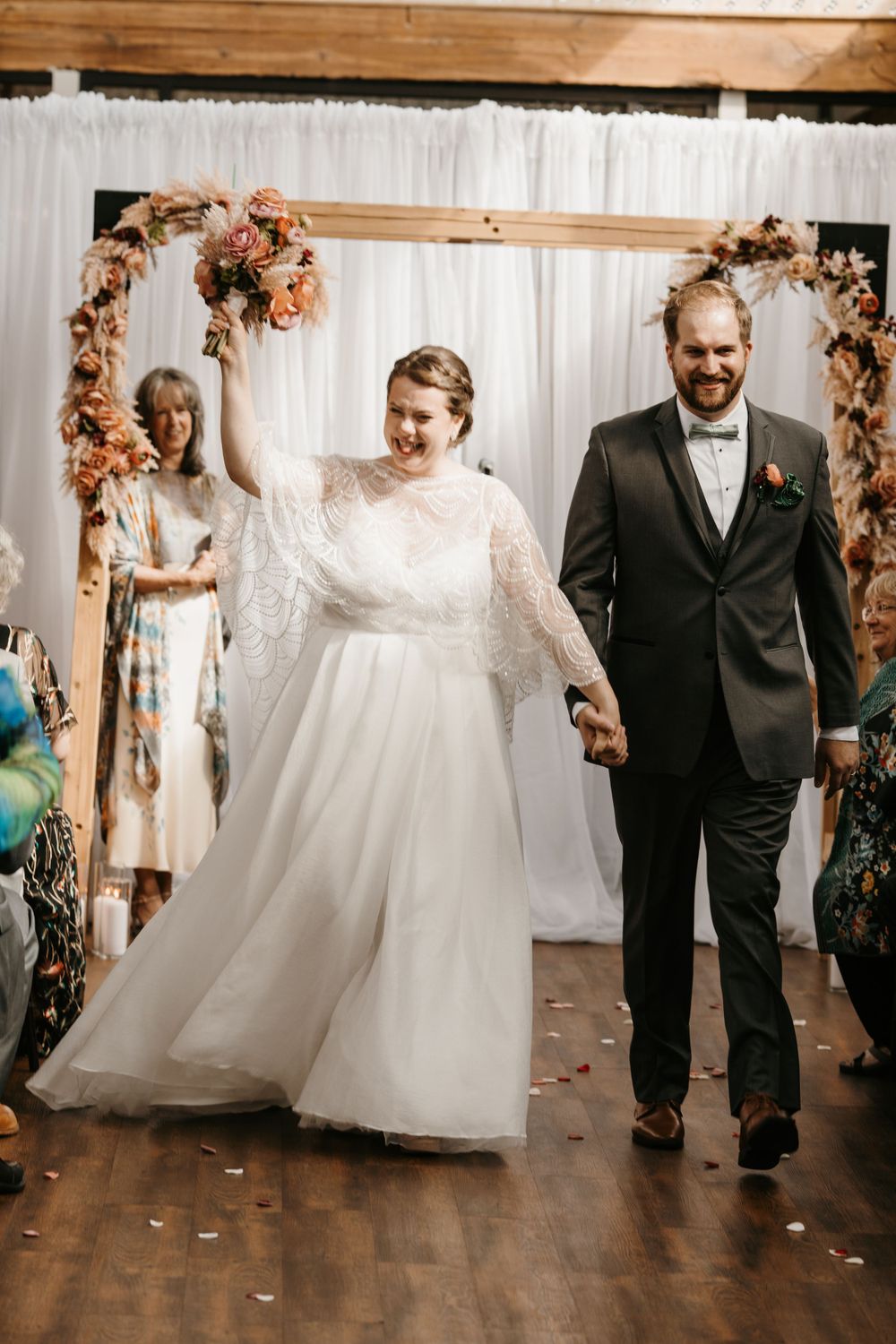 Newlyweds walk joyfully down the aisle as bride raises bouquet after indoor wedding ceremony.