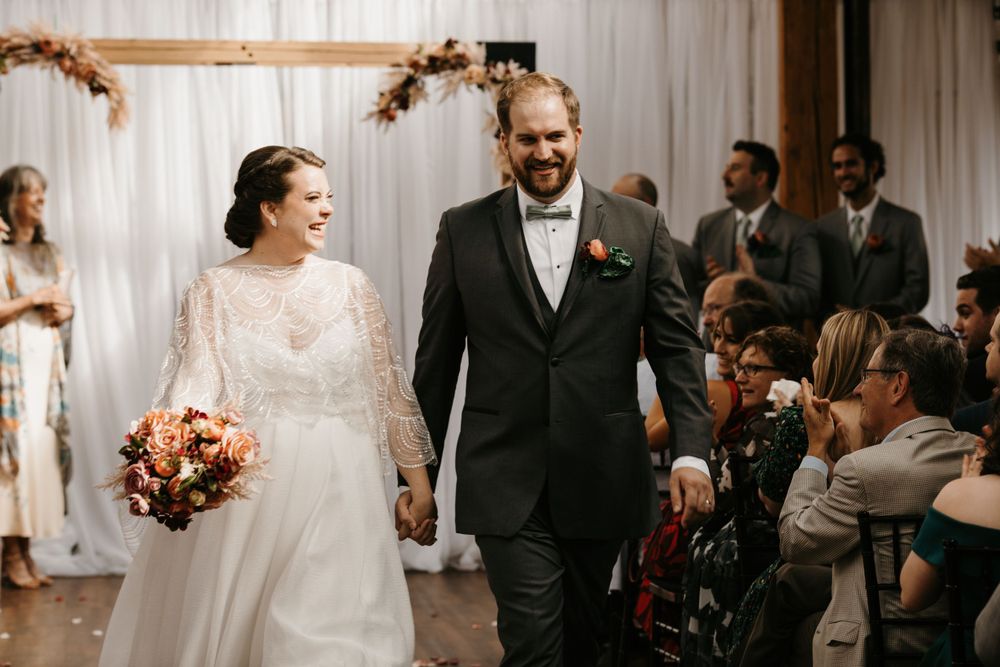 Smiling newlyweds walk hand-in-hand down the aisle as guests cheer during wedding recessional.