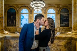 A romantic kiss under an ornate chandelier in a luxurious historic building with arched windows.
