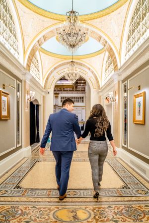 A couple walks hand in hand down an elegant hallway with chandeliers and ornate carpet.