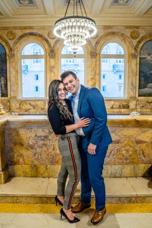 A happy couple poses in an ornate marble hall with chandelier and arched windows.