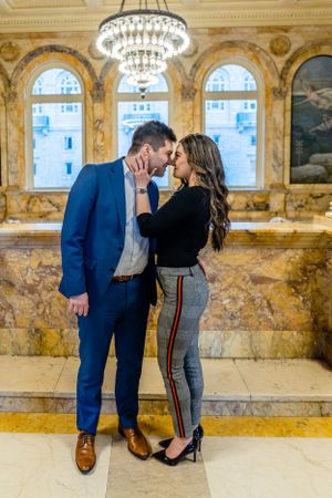 A couple shares an intimate moment in an elegant hall with crystal chandeliers and marble columns.