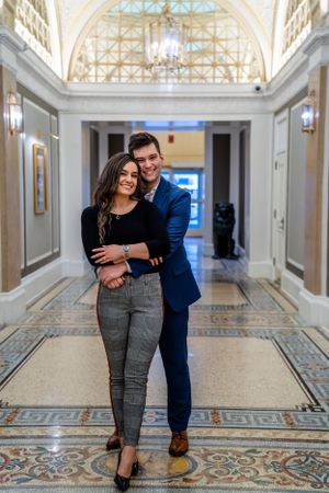 A couple embraces in an elegant hallway with ornate architectural details and a domed glass ceiling.