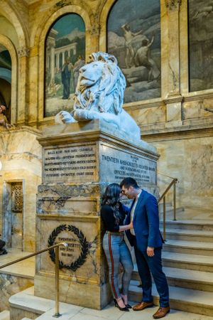 A couple embraces near a majestic stone lion statue in a grand historical building.