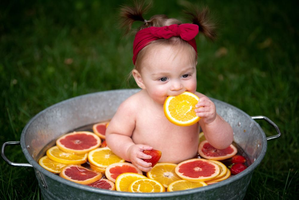 Baby in red bow sitting in metal tub with oranges and grapefruits during outdoor photo session.