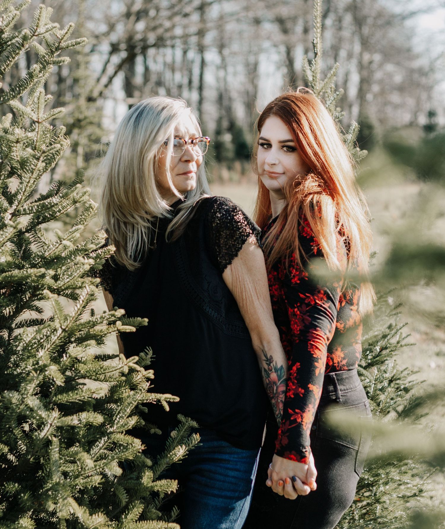 Two women posing together between evergreen trees in natural outdoor setting.